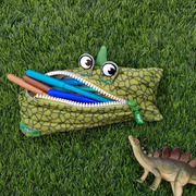 Green dinosaur-shaped pencil case with pens on grass, next to a toy dinosaur.
