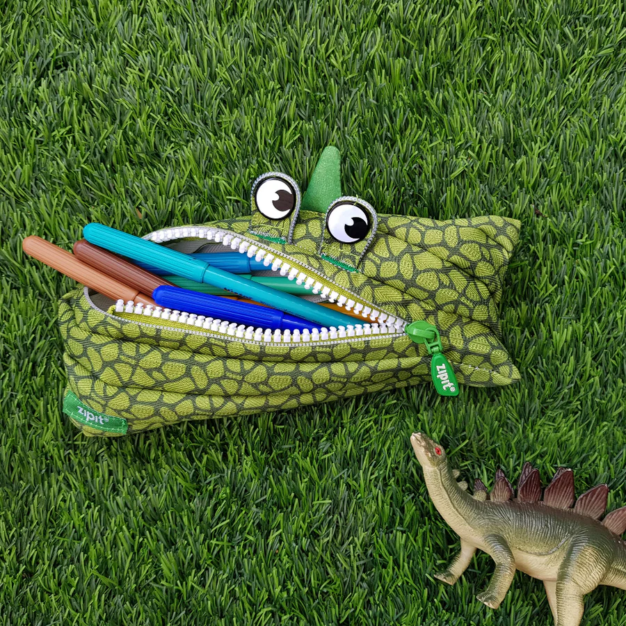 Green dinosaur-shaped pencil case with pens on grass, next to a toy dinosaur.