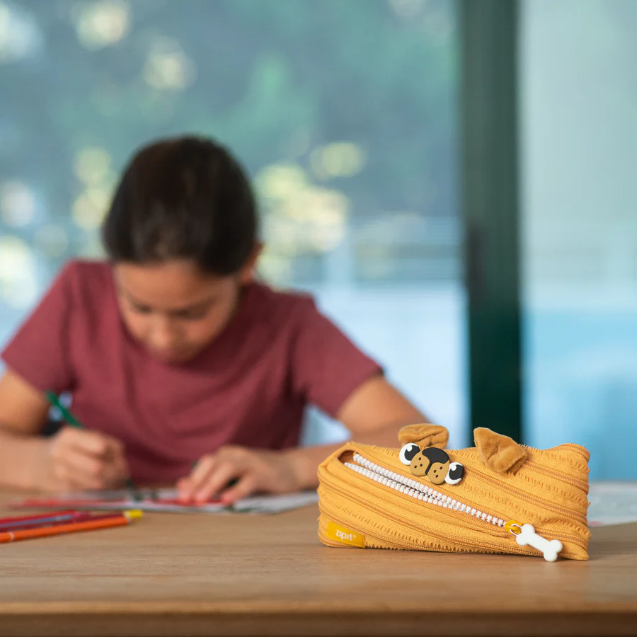 Dog-shaped pencil case on a table with a blurred child drawing in the background